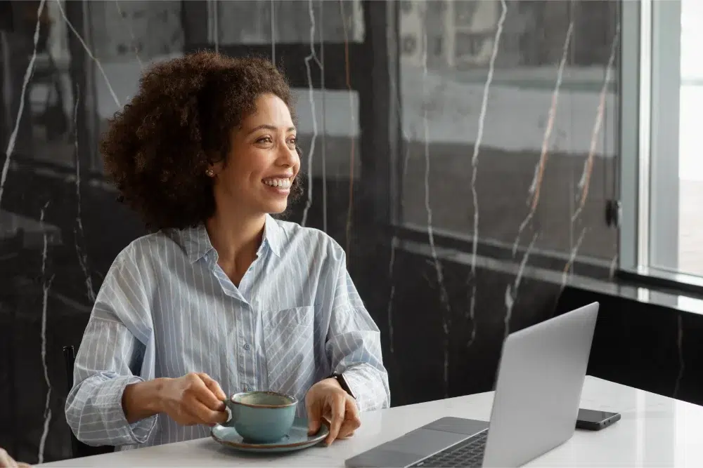 woman sitting at work desk with her morning coffee, smiling and feeling good at her job thanks to a good employee pension plan securing her future